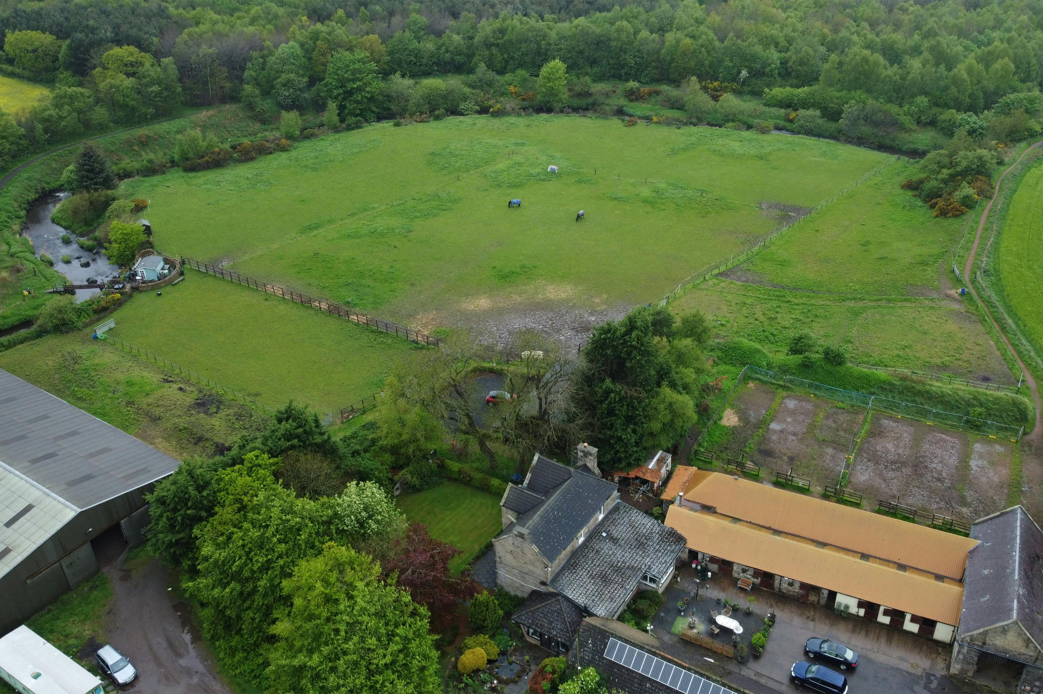 Aerial view of a green countryside landscape with open fields, stone walls, farm buildings, and surrounding woodland in the U