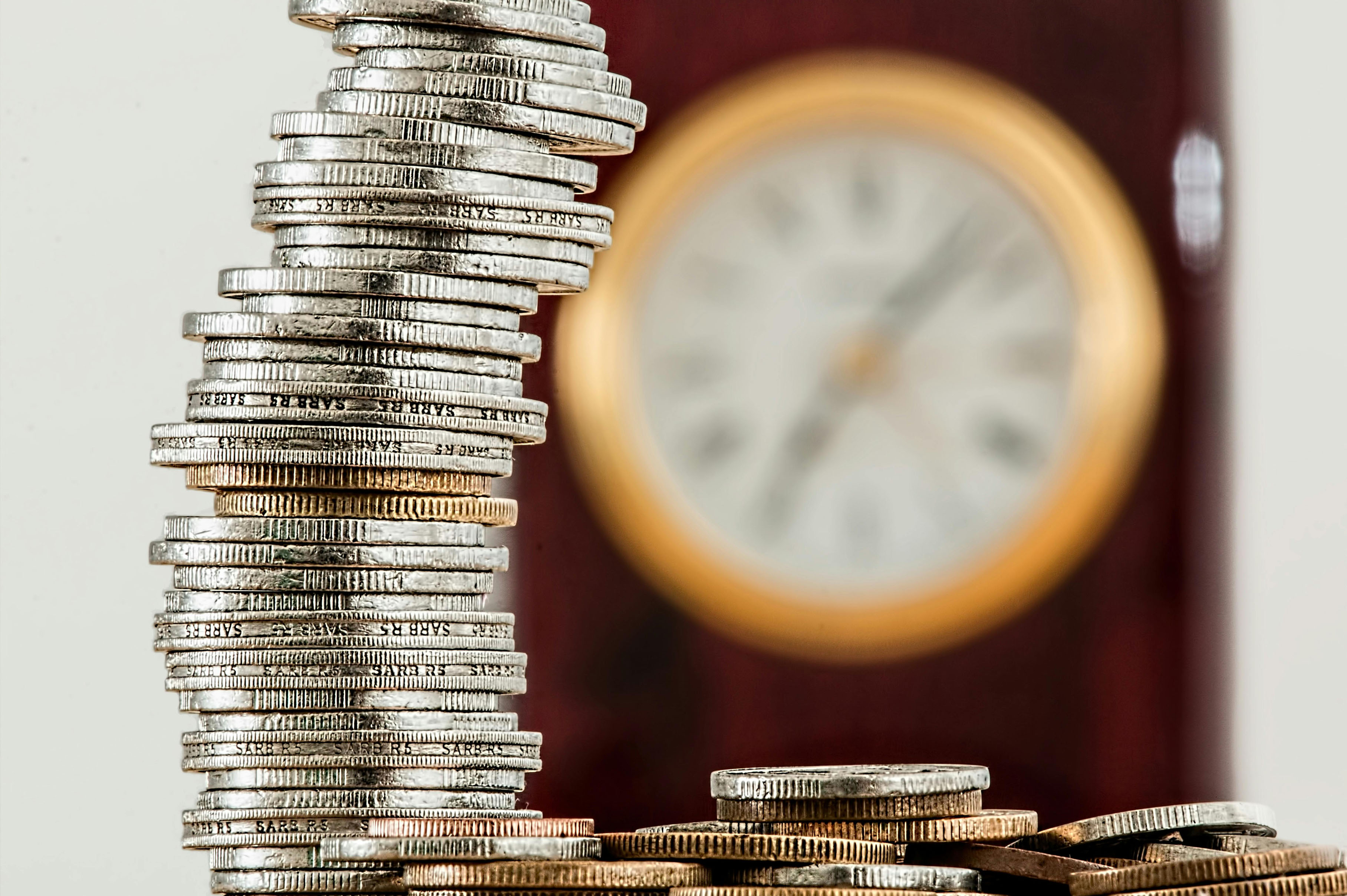 Stack of silver coins leaning in front of a blurred clock face, symbolising finance, investment and time value